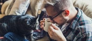 a man drinking a cup of coffee on the couch with his cat near him