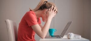 A stressed woman sitting at the table, staring at her laptop with her head in her hands. 