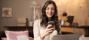 A smiling woman sitting on a couch with a laptop on her lap and a phone in her hand.