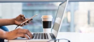 a woman holding her phone over her laptop next tu a cup of coffee and a pair of glasses